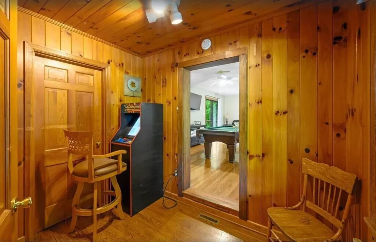 An interior view of a corner with nostalgic wood paneling, a classic arcade game console set beside a doorway, and a bar stool. The setup evokes playful competition and retro charm, ideal for family fun and memorable showdowns.
