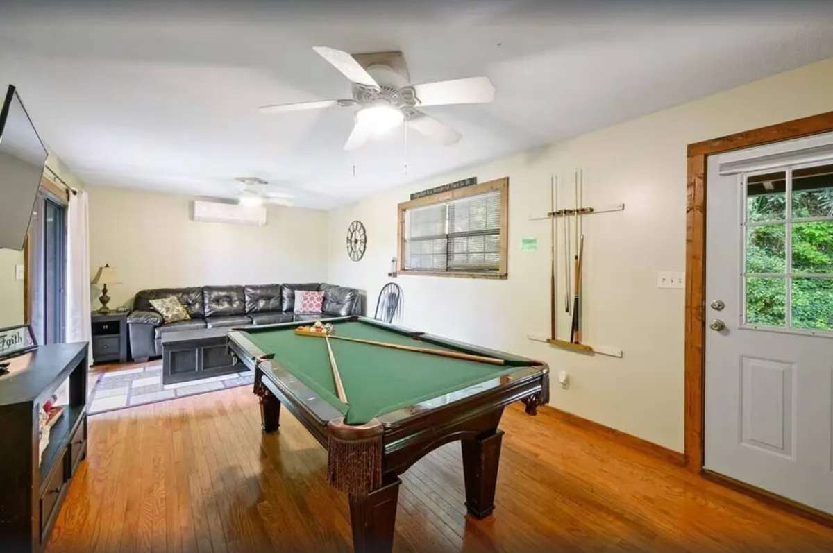 A spacious game room with a pool table, leather sofa, and a large TV, featuring a door that leads outside. The comfortable arrangement encourages both spirited competition over pool and cozy movie nights, blending relaxation and excitement.