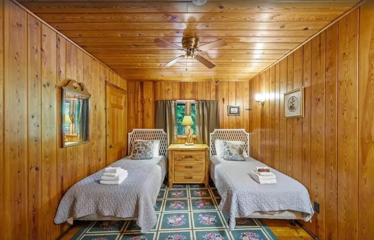 A cozy children's play area with two twin beds, a nightstand, a mirror, and a window with curtains. The setup is warm and inviting, providing a safe, kid-friendly corner for creative play, rest, or quiet reading.