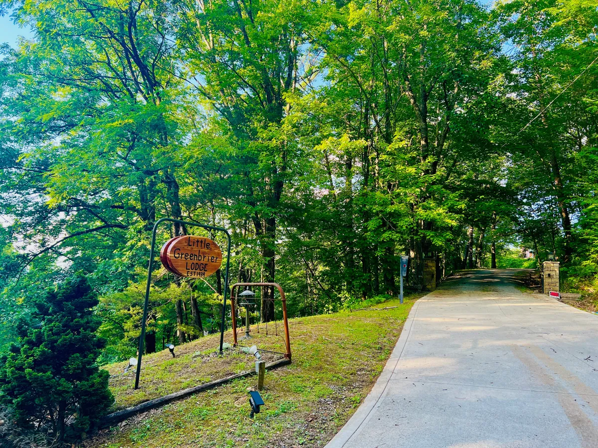 An inviting exterior shot of Little Greenbrier Lodge, featuring a welcoming sign, mature trees, and a pathway that draws guests toward the lodge. The natural setting hints at both winter and summer activities, with ample outdoor space for gathering and relaxation.