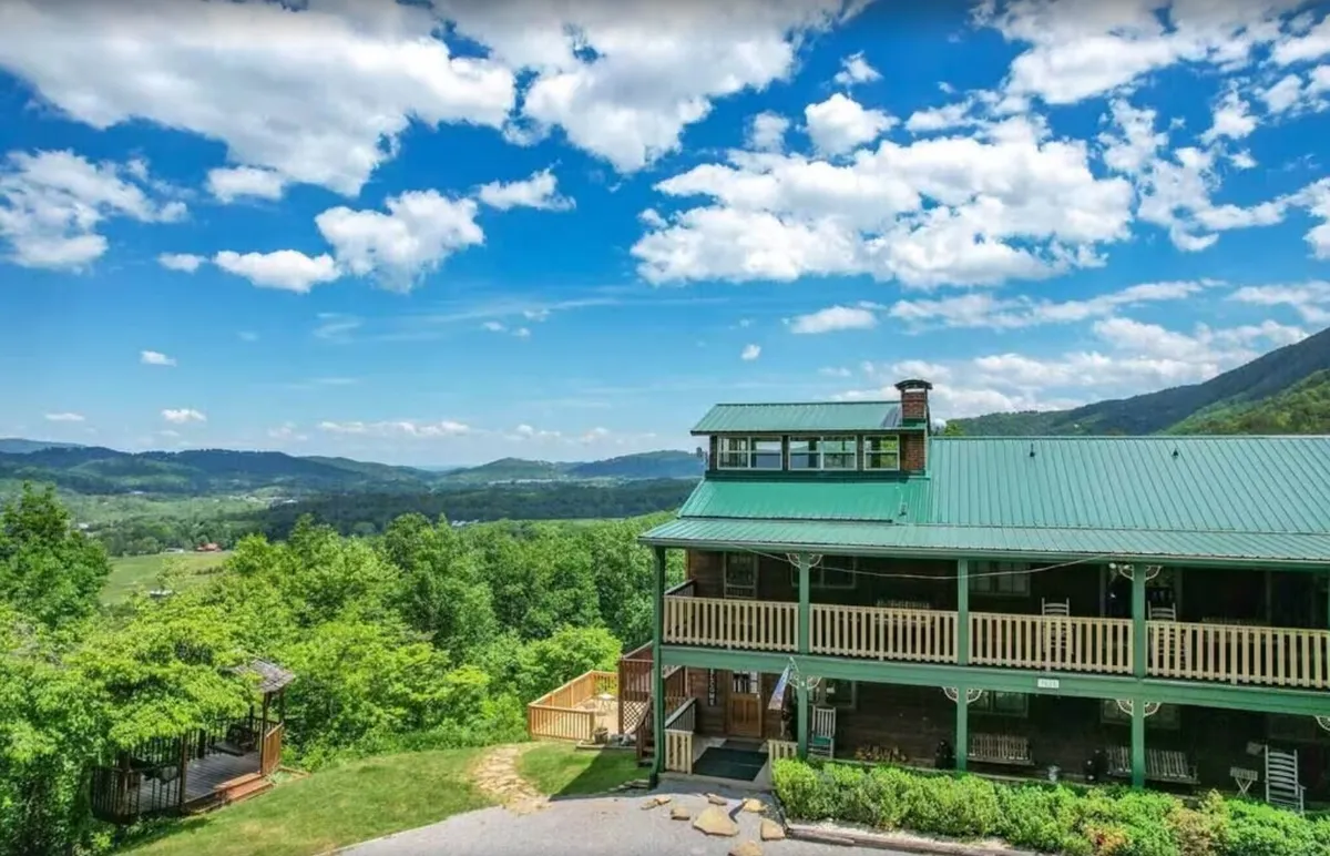 This image showcases the house with a green roof set against an expansive mountain view, with a welcoming porch area in the foreground. The tranquil setting highlights lush greenery and the vastness of the Smoky Mountains beyond, setting a perfect scene for outdoor relaxation or preparation before a day in the park.
