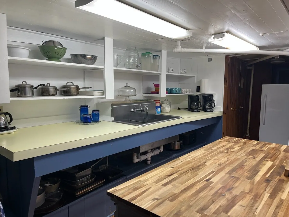 The image captures a kitchen island with shelves full of cookware, a sink for food prep, and multiple coffee makers. Wood accents and practical design elements create a homely yet semi-commercial feel, ideal for group cooking and interactive meal prep.