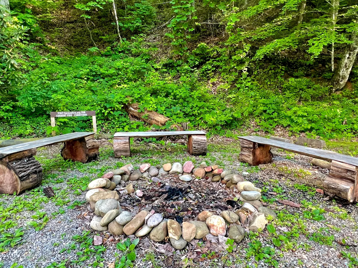 A classic fire pit is surrounded by wooden benches and encircled by a serene, green landscape. The arrangement evokes the warmth of gathering for campfire stories, stargazing, and roasting marshmallows with friends or family.