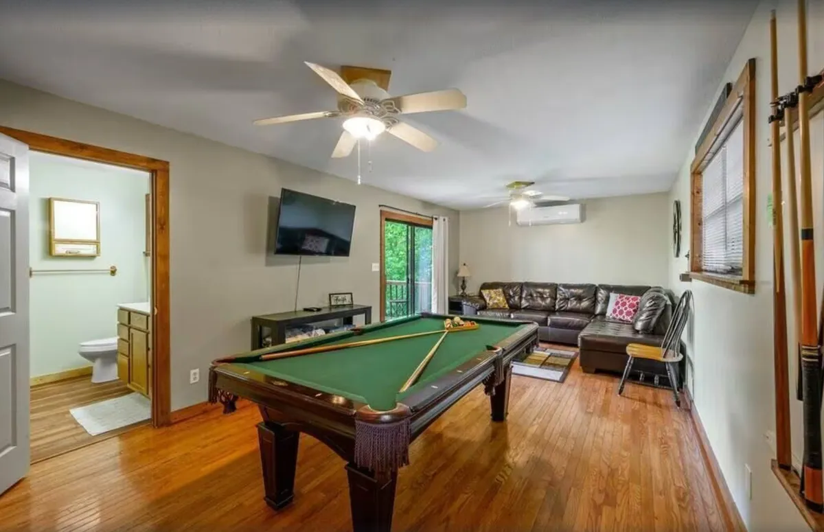 A vibrant interior game room featuring a regulation pool table at the center, a plush leather sofa for comfortable seating, and a flat-screen television mounted on the wall. The wood-paneled flooring and cozy arrangement create an inviting atmosphere, perfect for group gatherings and friendly competition.