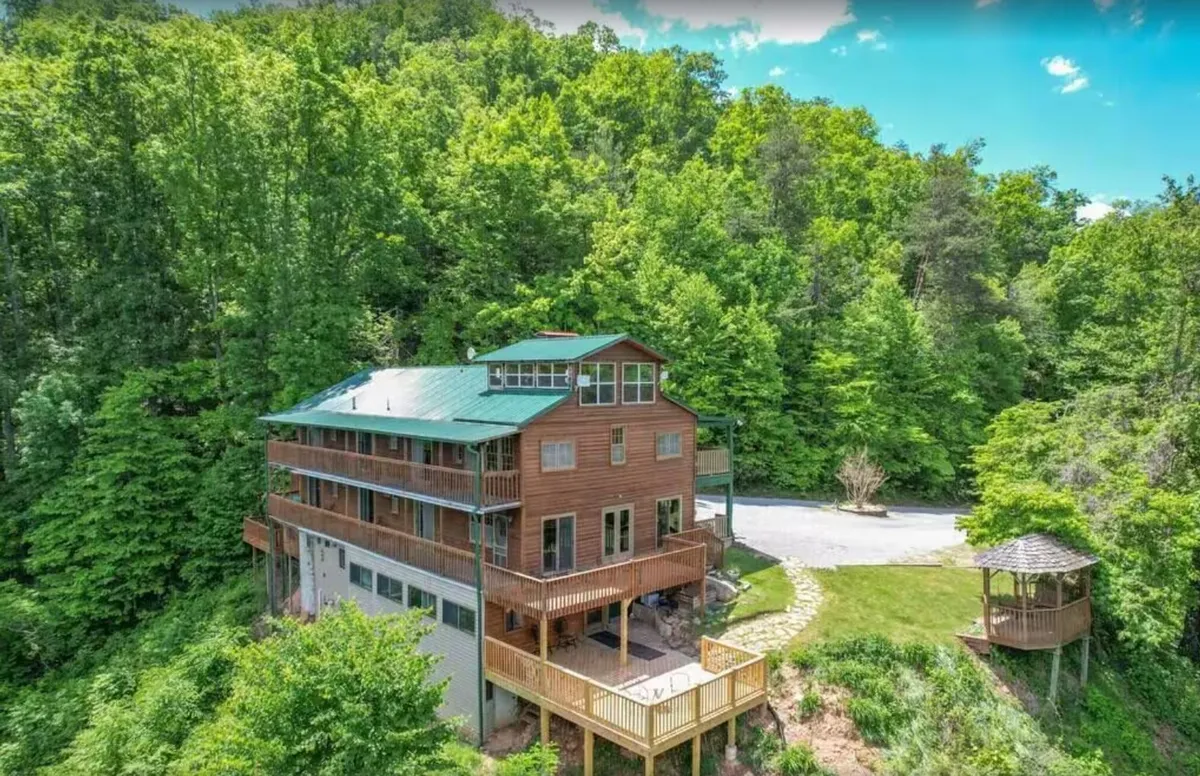 This image displays the wooden exterior of the lodge, nestled among a green canopy of trees, with an expansive outdoor deck and a gazebo visible. The inviting setup emphasizes spaces ideal for outdoor gatherings and group activities, offering both connection with nature and ample room for celebrations.