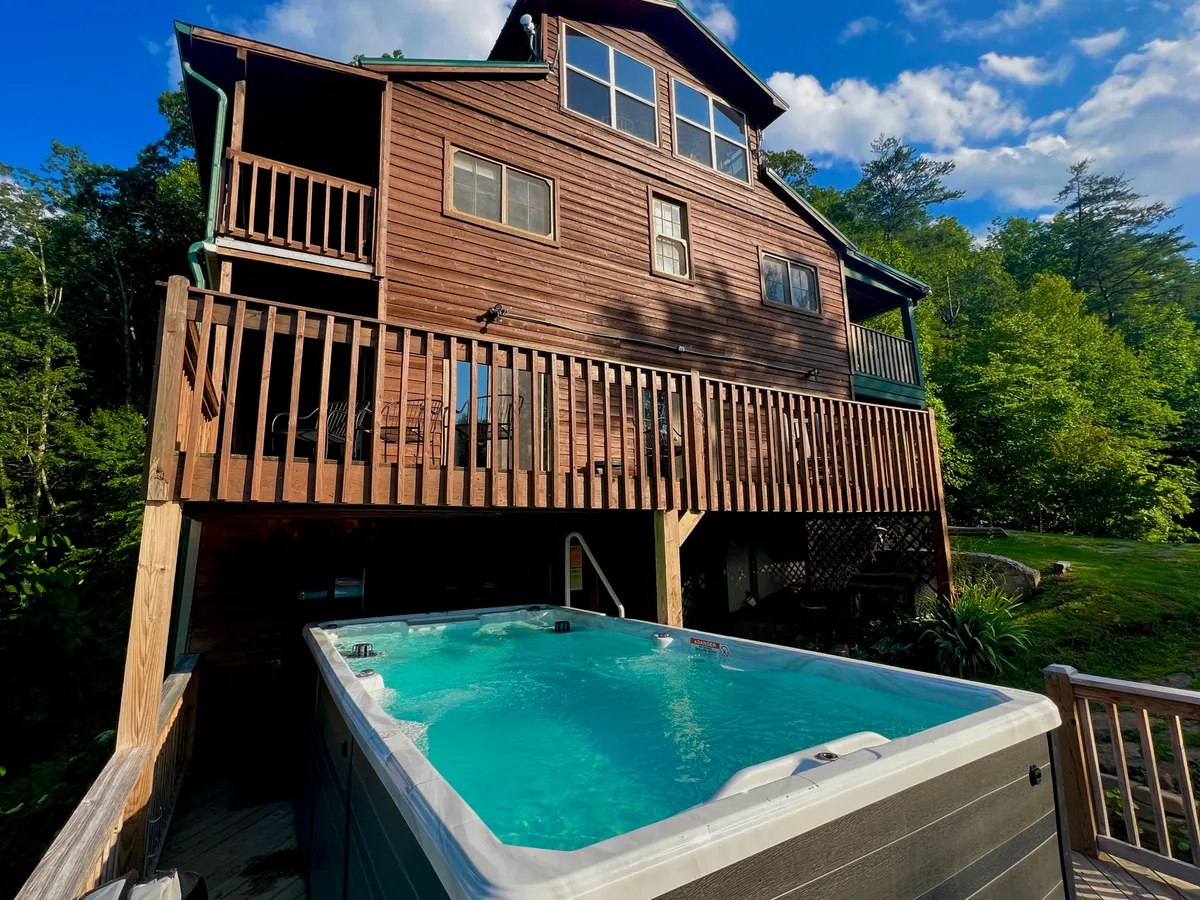 This image shows a wooden deck with a hot tub, the house facade, and surrounding trees. The setup is inviting, with space for outdoor meals and group relaxation, illustrating additional amenities that foster togetherness and celebration.
