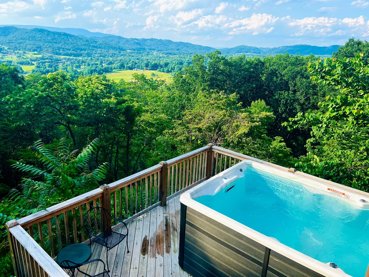 A wooden deck features a hot tub overlooking panoramic mountain views, framed by forested hills and open sky. The inviting scene suggests relaxation, with opportunities for soaking in the scenery at sunset or stargazing after dark.