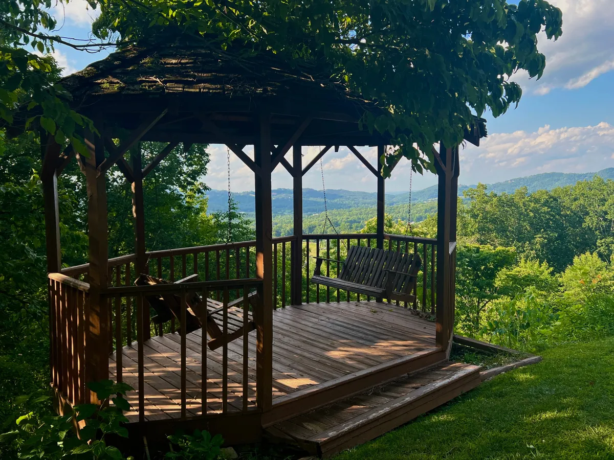 The image captures a wooden gazebo with swing seats set against the backdrop of distant mountains and lush greenery. The gazebo provides a focal point for outdoor socializing, relaxation, and small group gatherings surrounded by natural beauty.