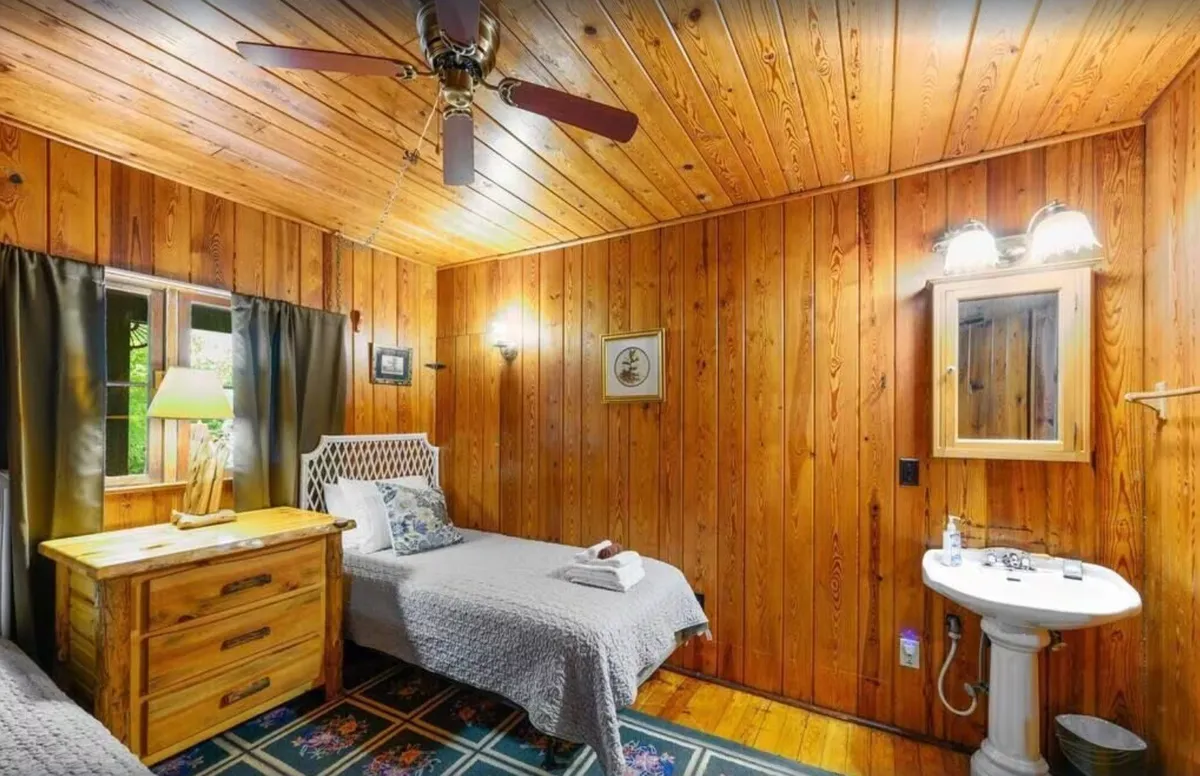 A bedroom with two beds, wood paneling, a nightstand lamp, and a vanity with a mirror. The room is inviting and functional, offering both personal space and shared accommodation options for various guest groupings.