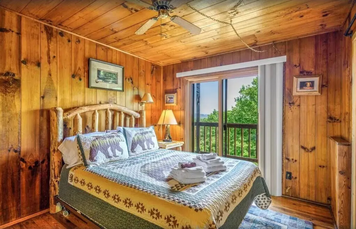 A warmly lit bedroom with a wooden bed frame, colorful quilt, and a large window offering a scenic view. The space feels rustic but comfortable, with natural light, inviting bedding, and serene outdoor sights that suggest a tranquil retreat.