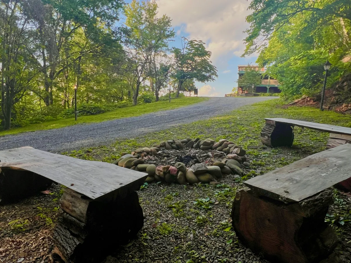 A cozy fire pit area encircled by wooden benches sits amidst trees and gravel paths, with a distant building visible through the foliage. The space is designed for communal gatherings, storytelling, and relaxing after a day of adventure, embodying the spirit of outdoor camaraderie.